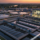 صورة قريبة لمنشأة صناعية تستخدم أنواع معالجة المياه المتقدمة، تظهر فيها أحواض الترسيب الأولية والمبنى الإداري للمحطة بتصميم زجاجي عصري أثناء وقت الشفق.