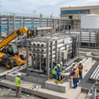 فريق فني يقوم بربط التوصيلات النهائية لوحدات معالجة المياه المعقدة، مع ظهور رافعة شوكية (JCB) وخزانات ضخمة تحت إشراف أفضل شركة تركيب محطات تحلية.